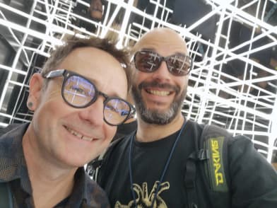 Zwei Mitarbeitende stehen vor einer Lichtinstallation und lachen in die Kamera.